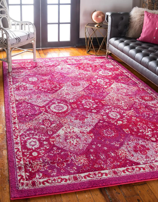 Magenta patterned rug set on a hardwood living room floor with a black leather couth and white wicker chair.