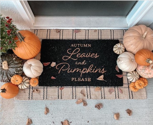 Festive mat set in front of a door that reads "autumn leaves and pumpkins, please", surrounded by several pumpkins on either side