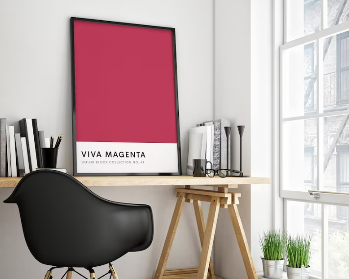 A framed Viva Magenta poster set on a desk, next to a window.