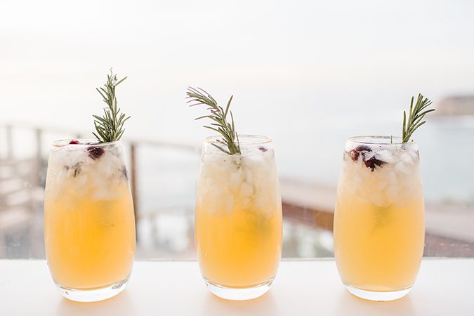Three cocktail glasses with a bright yellow drink inside, topped with cranberries and a sprig of rosemary.