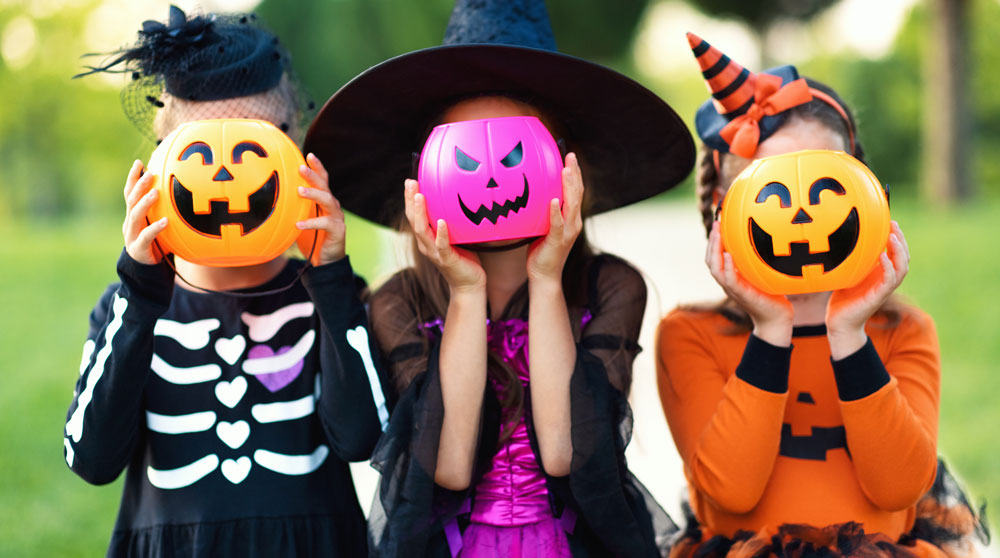 Three kids in Halloween costumes, holding trick-or-treat pumpkin buckets in front of their faces