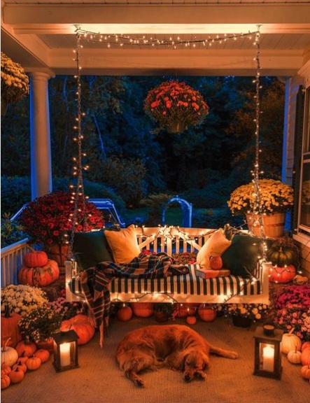 A front porch decked out in pumpkins and mum with a porch wing that's decorated with string lights, pillows, and a blanket