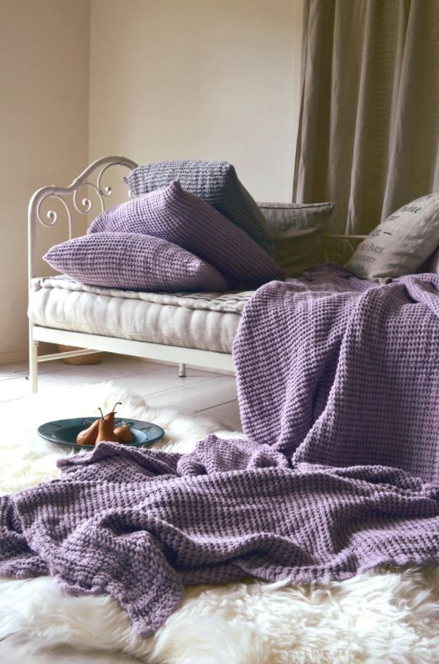 Lilac knit blanket hanging over a beige colored bed.