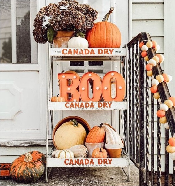A repurposed Canada Dry shelf set on a porch and stacked with pumpkins, pots, and marquee "B-O-O" lights