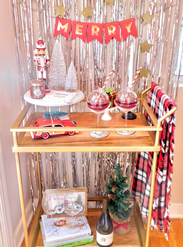 Bar cart set in front of a festive tinsel backdrop with a red banner that says Merry and hanging gold stars.