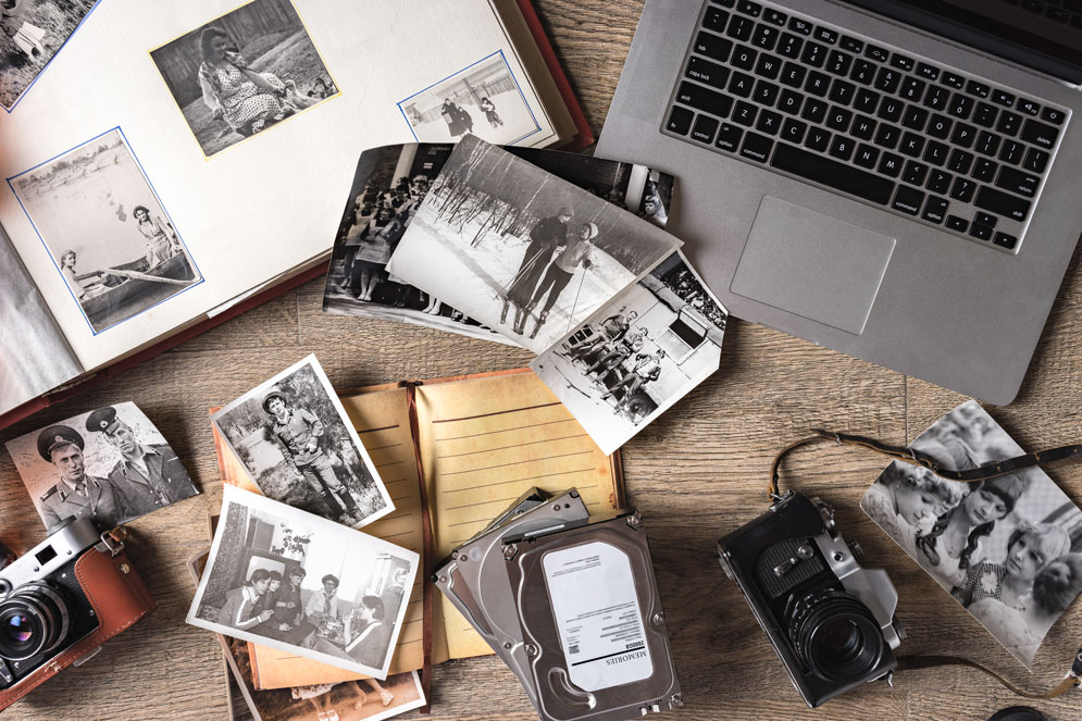 Vintage family photos and album laying flat on a wooden background with a film camera, old notepad and modern laptop.