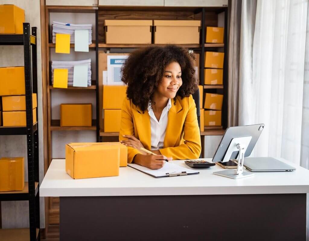 Young female employee sitting at desk writing while looking at laptop screen with empty boxes behind her.