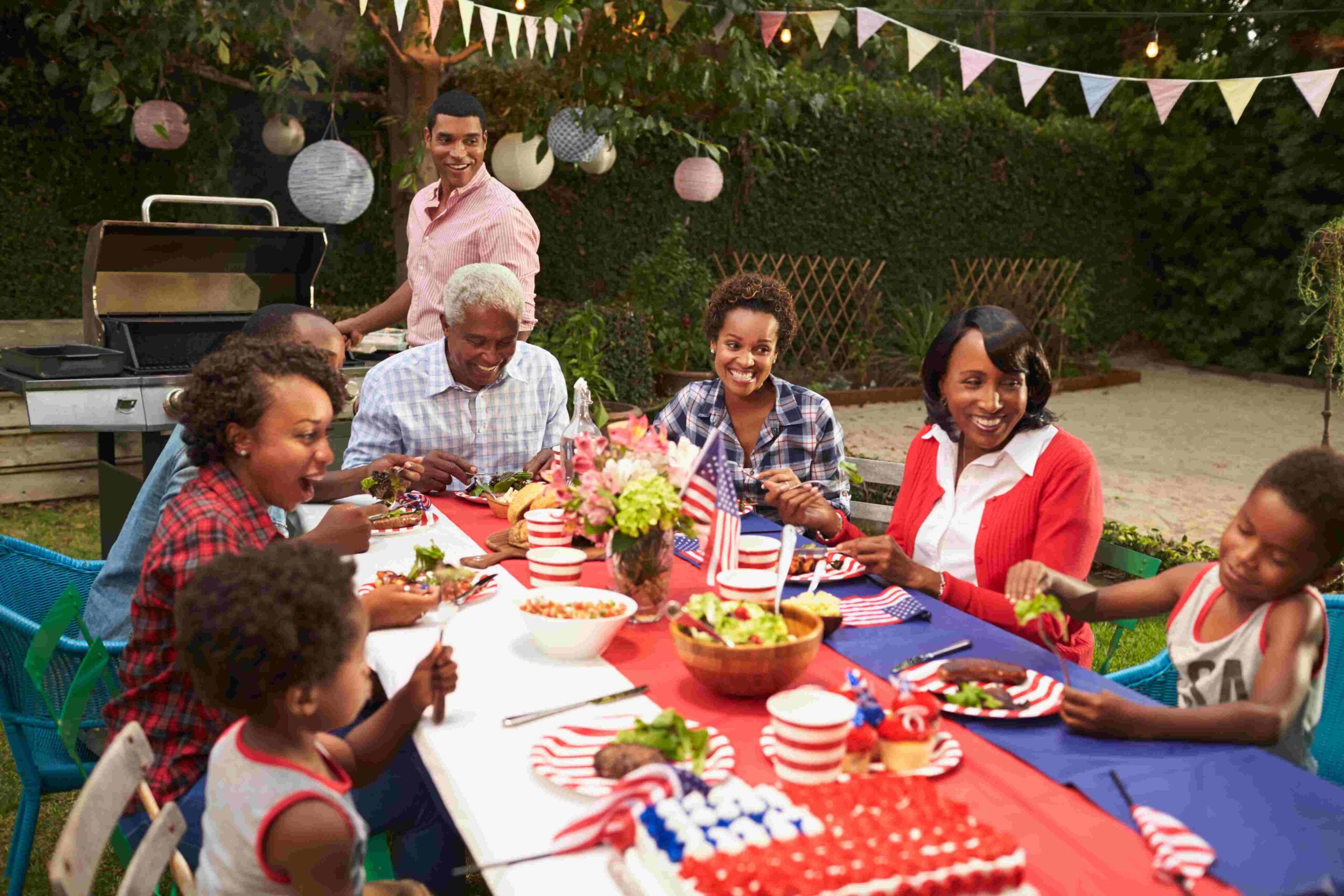 Family celebrating the Fourth of July with a backyard barbecue, patriotic decorations, and festive red, white, and blue table settings.